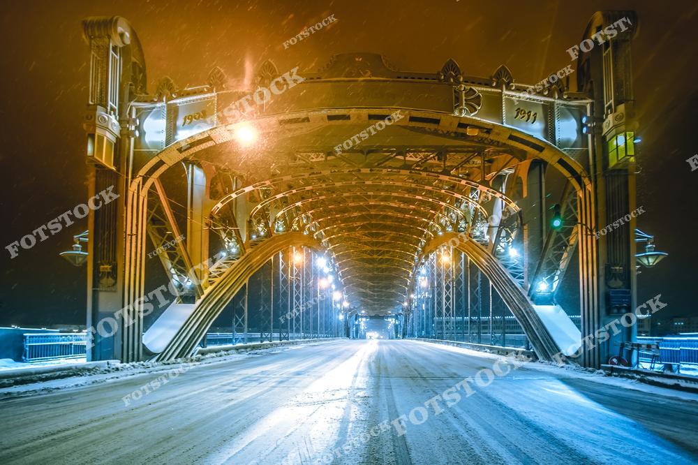 На фотографии изображён Большеохтинский мост, покрытый лёгким снегом в зимнюю ночь. Металлические арки моста создают гармоничную композицию, а уличные огни добавляют тепла в холодный пейзаж. Снежные сугробы и освещённые конструкции подчеркивают величие инженерного решения. Этот мост — одна из знаковых достопримечательностей Санкт-Петербурга, соединяющая его исторические районы.